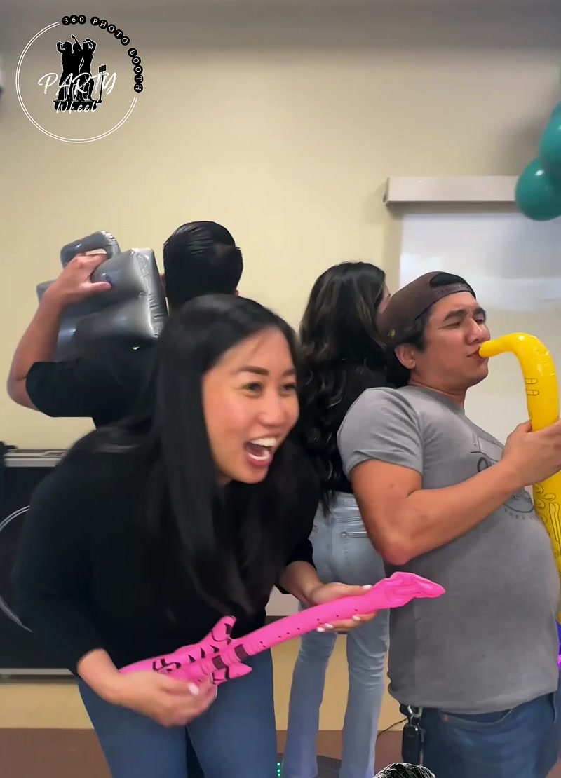 Smiling woman playing a pink inflatable guitar while a man in a gray shirt plays a yellow inflatable saxophone at a party