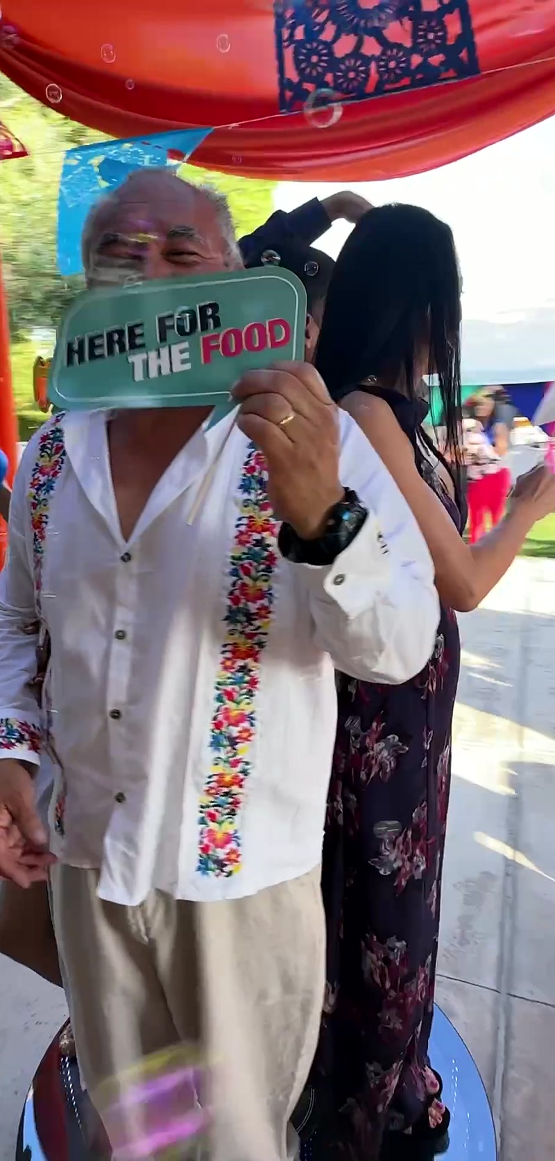 Older man holding a Here for the Food sign at an outdoor party with a woman behind him