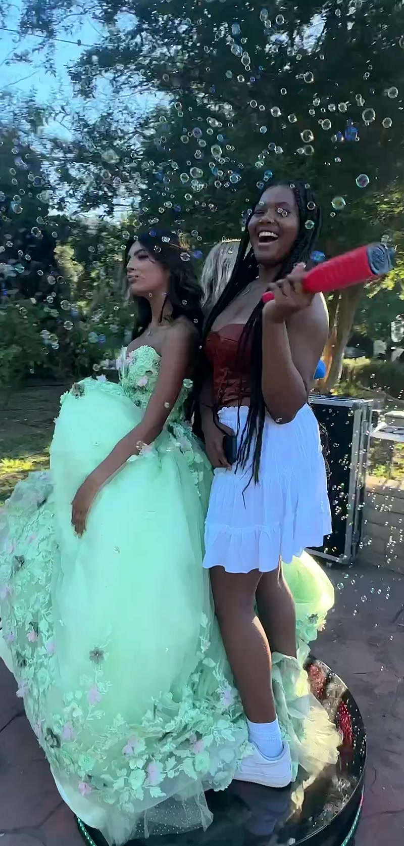 Two young women laughing in a shower of bubbles outdoors, one holding a bubble blaster